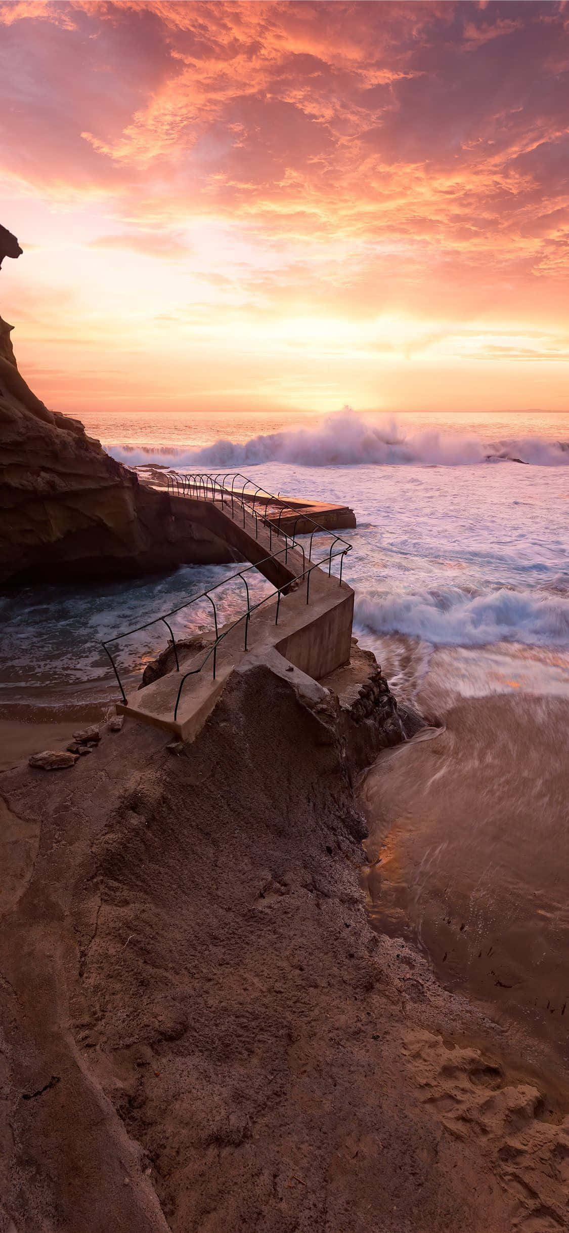 Coastal Sunset 桌布 壁紙 — 戲劇性的日落將天空染成火熱的橙色和粉紅色，海浪拍打著崎嶇的海岸懸崖，一條混凝土樓梯通向海洋。