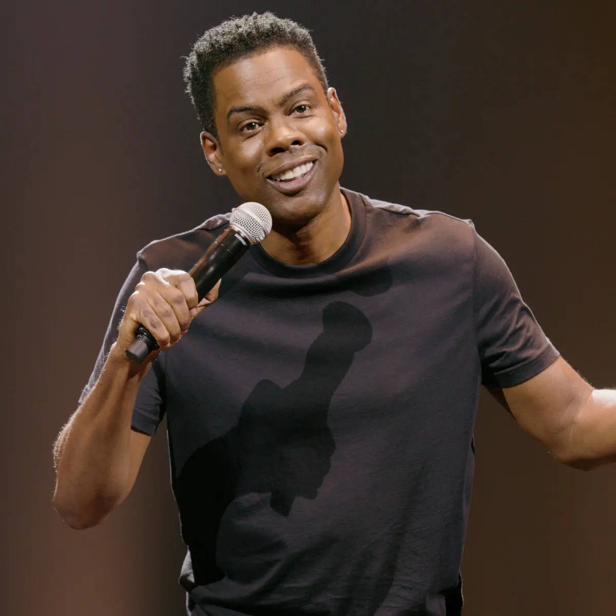 克里斯·洛克動態演出 壁紙 — 喜劇演員基斯·洛克在溫暖的聚光燈下在舞台上拿著麥克風微笑。