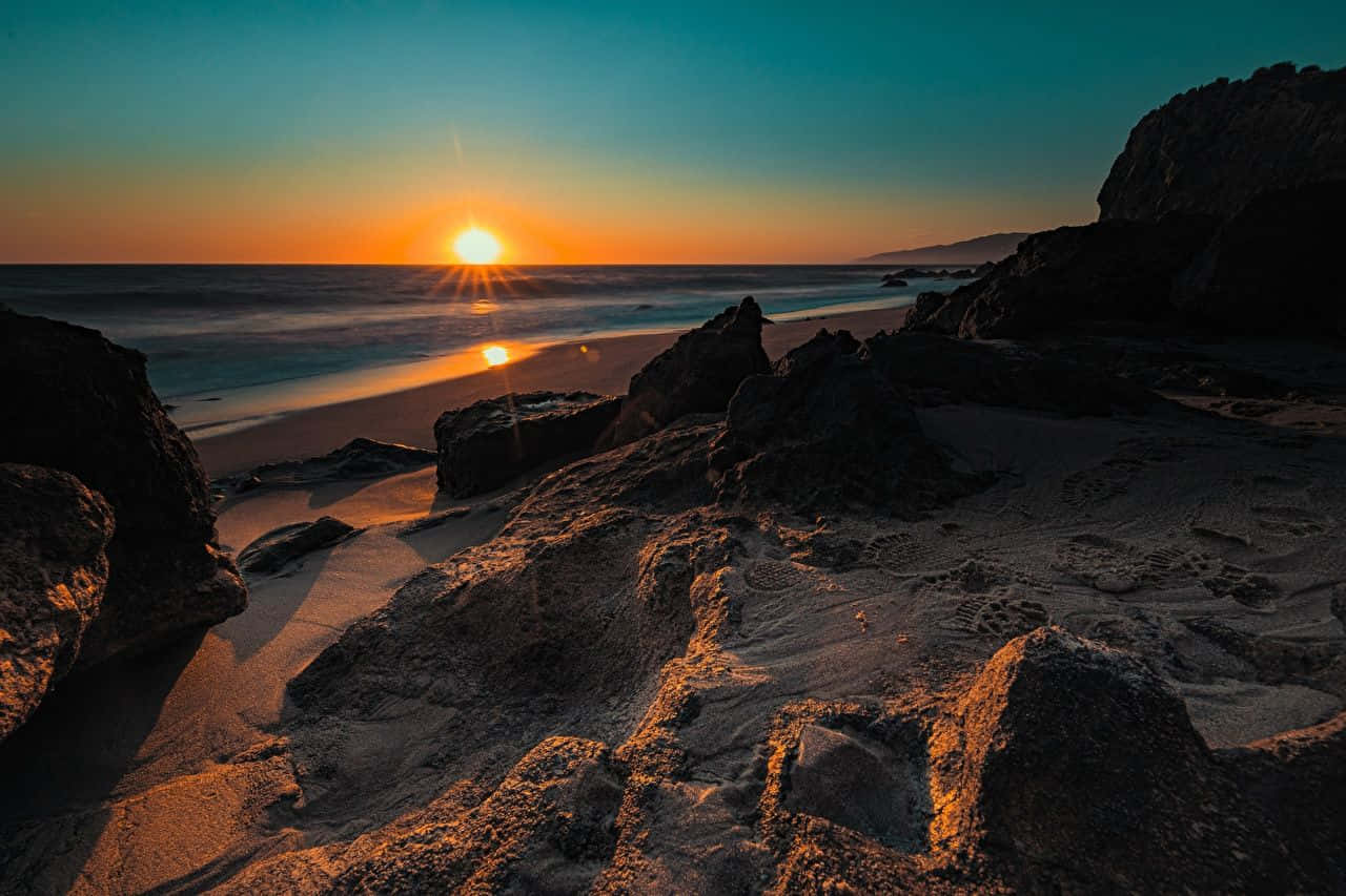 Coastal Sunset 桌布 壁紙 — 夕陽將金色光芒灑在崎嶇的岩石和沙灘上，海浪在漸變的天空下輕輕拍打。