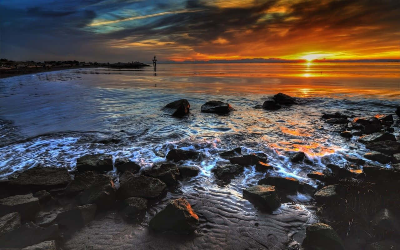 Coastal Sunset 桌布 壁紙 — 夕陽將天空染成火橙色和深藍色，海浪拍打著寧靜海岸線上的黑色岩石。