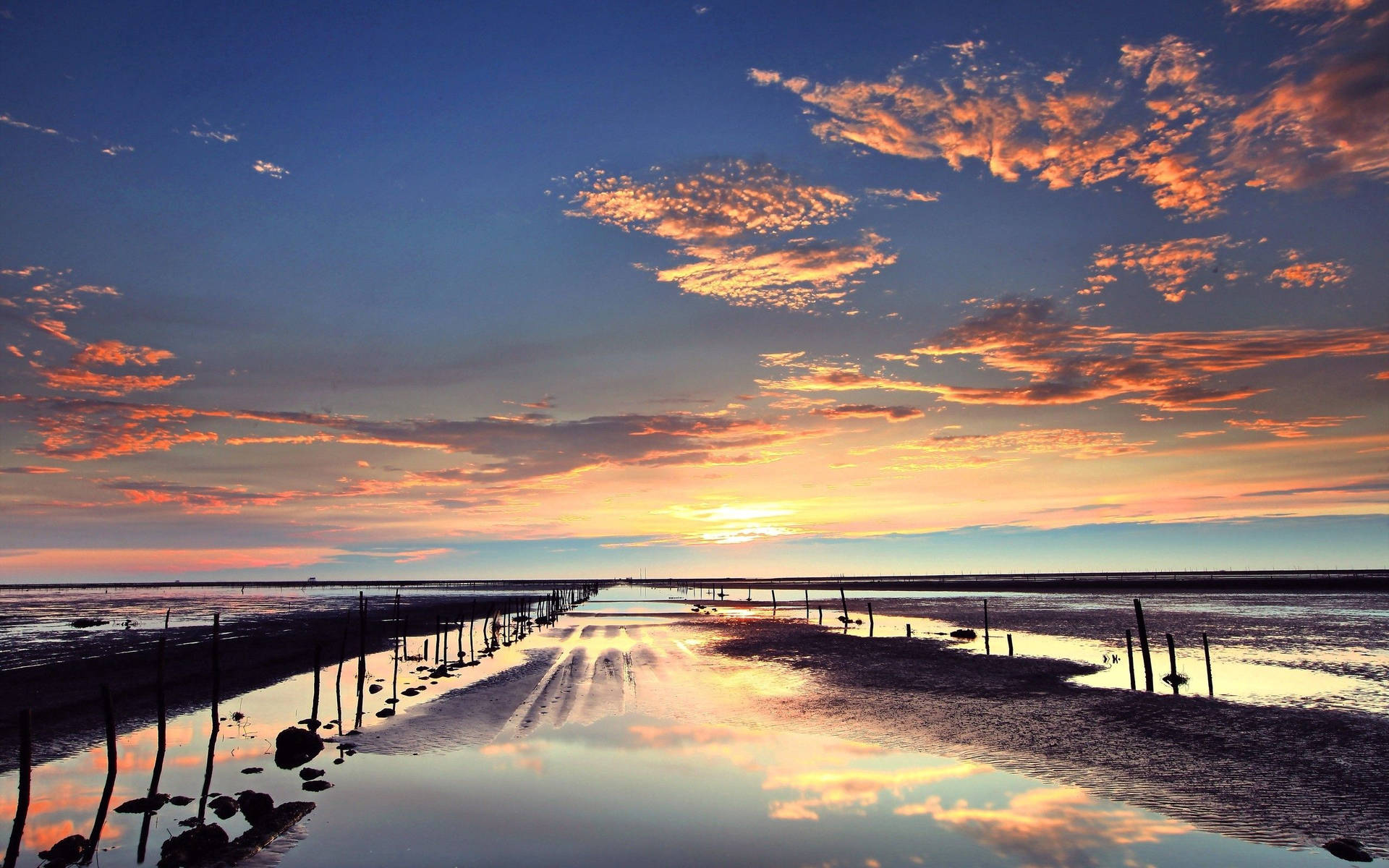 海與夕陽自然風光 壁紙 — 夕陽將天空染成火橙色和深藍色，雲朵倒映在泥濘潮灘旁的平靜水面上。