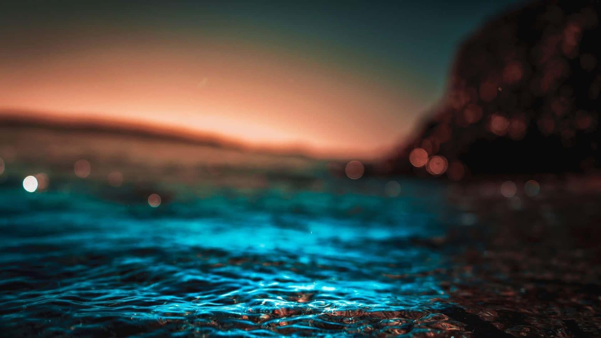  壁紙 — 夕陽將天空染成溫暖的橙黃色調，映照在漣漪的碧綠水面上，背景是模糊的岩石峭壁。