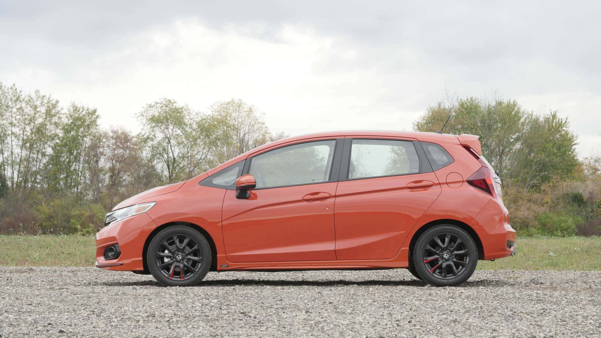 Honda Fit 桌布 壁紙 — 一輛鮮橙色的本田Fit配黑色輪圈，停泊在碎石上，旁邊有一排樹木，天空多雲。