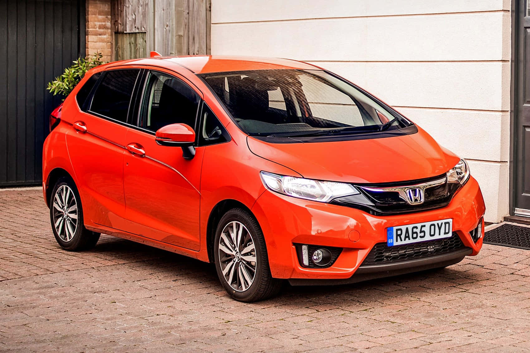 Honda Fit 桌布 壁紙 — 一輛鮮橙色的本田 Jazz 停泊在磚砌車道上，旁邊是一棟帶木質裝飾的現代房屋。