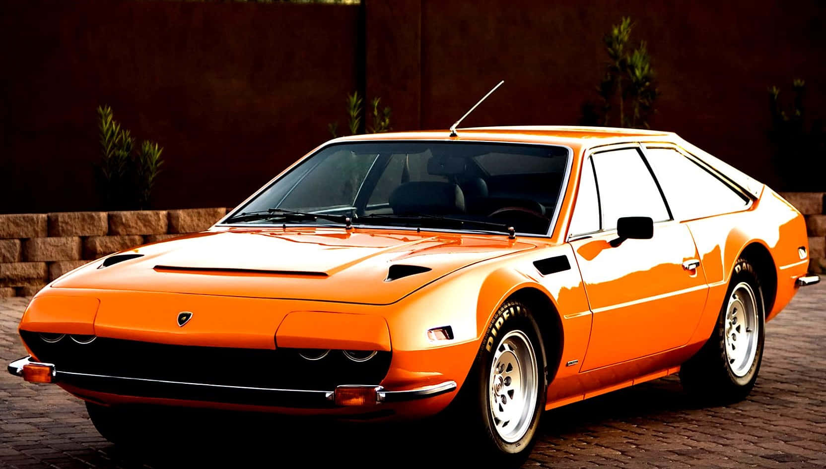 Lamborghini Silhouette 桌布 壁紙 — 一輛鮮橙色的經典林寶堅尼Countach停泊在磚砌車道上，背景有石牆和綠植。