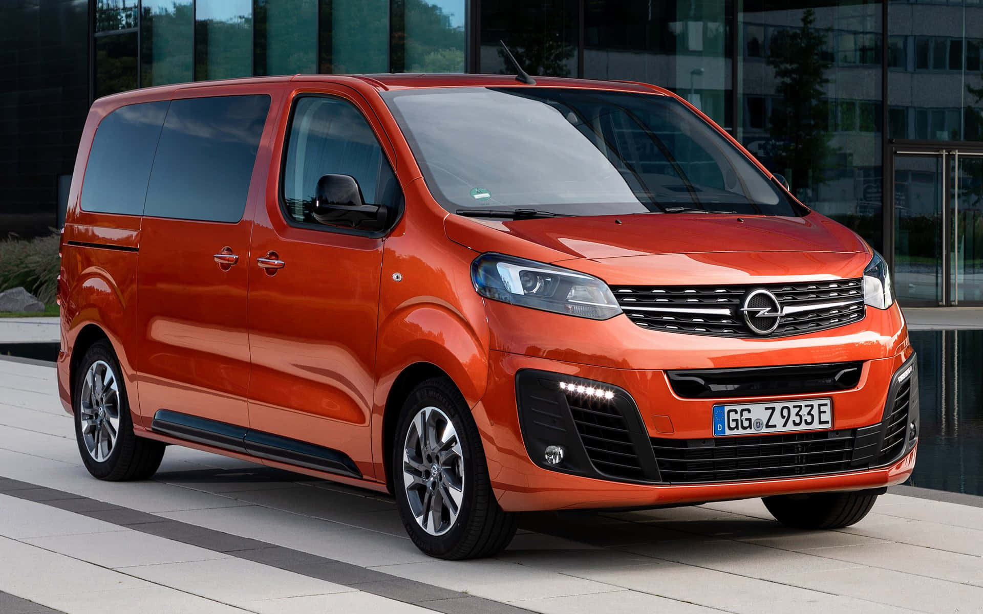 Opel Zafira 桌布 壁紙 — 一輛鮮橙色的歐寶Zafira Tourer時尚地停放在現代瓷磚廣場上，旁邊是一座玻璃建築。