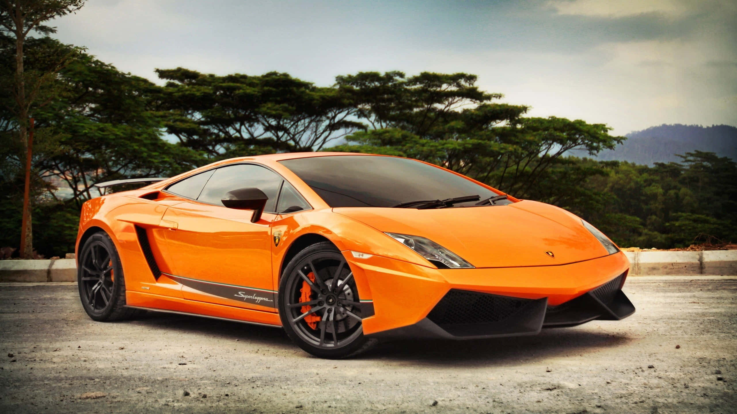 Lamborghini Gallardo 桌布 壁紙 — 一輛鮮橙色林寶堅尼Gallardo Superleggera停泊在碎石路上，周圍有茂密的綠樹和遠山，天空多雲。