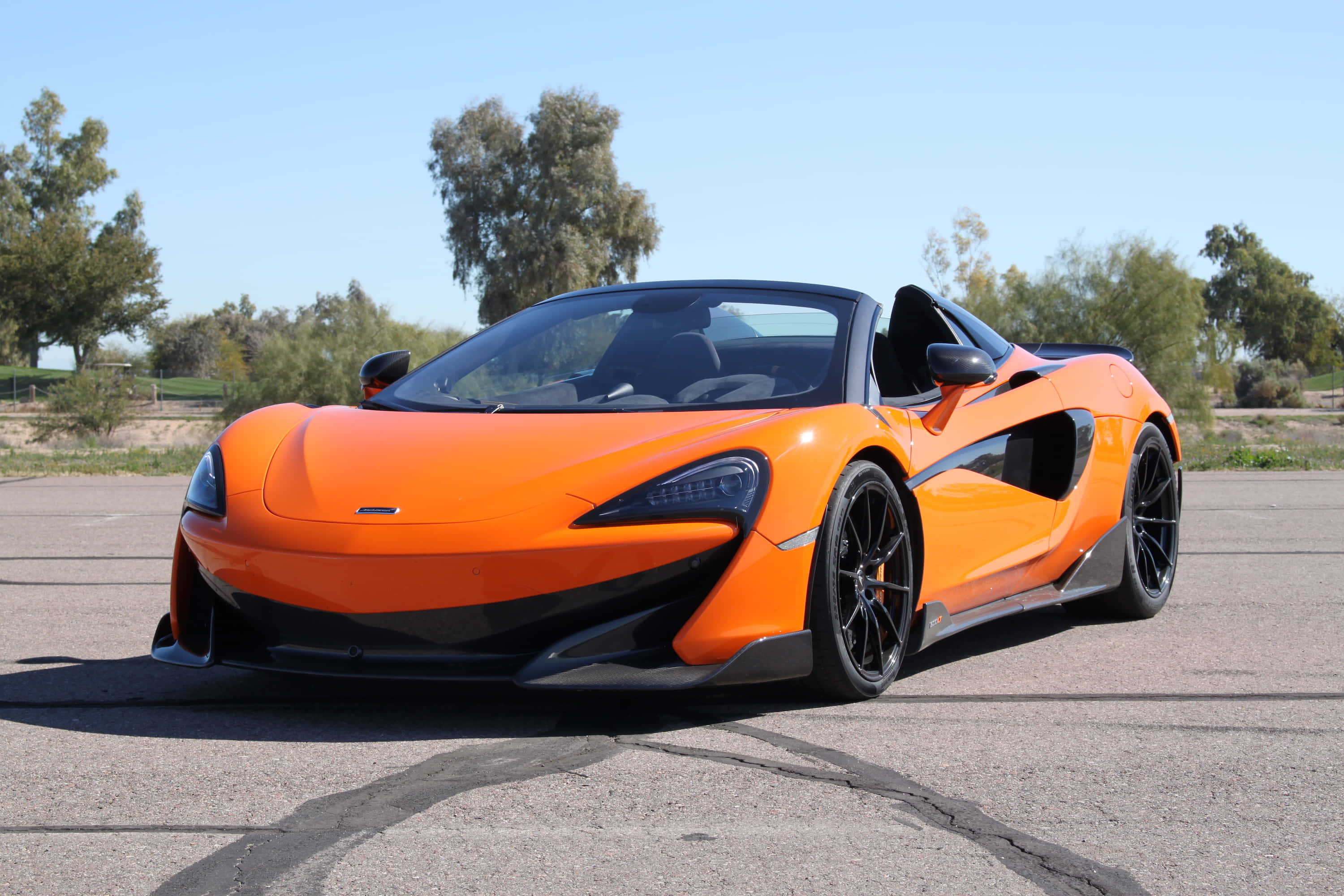 Mclaren 600lt Spider 桌布 壁紙 — 一輛鮮橙色的McLaren 570S Spider停泊在裂開的瀝青路上，背景是清澈的藍天和樹木。
