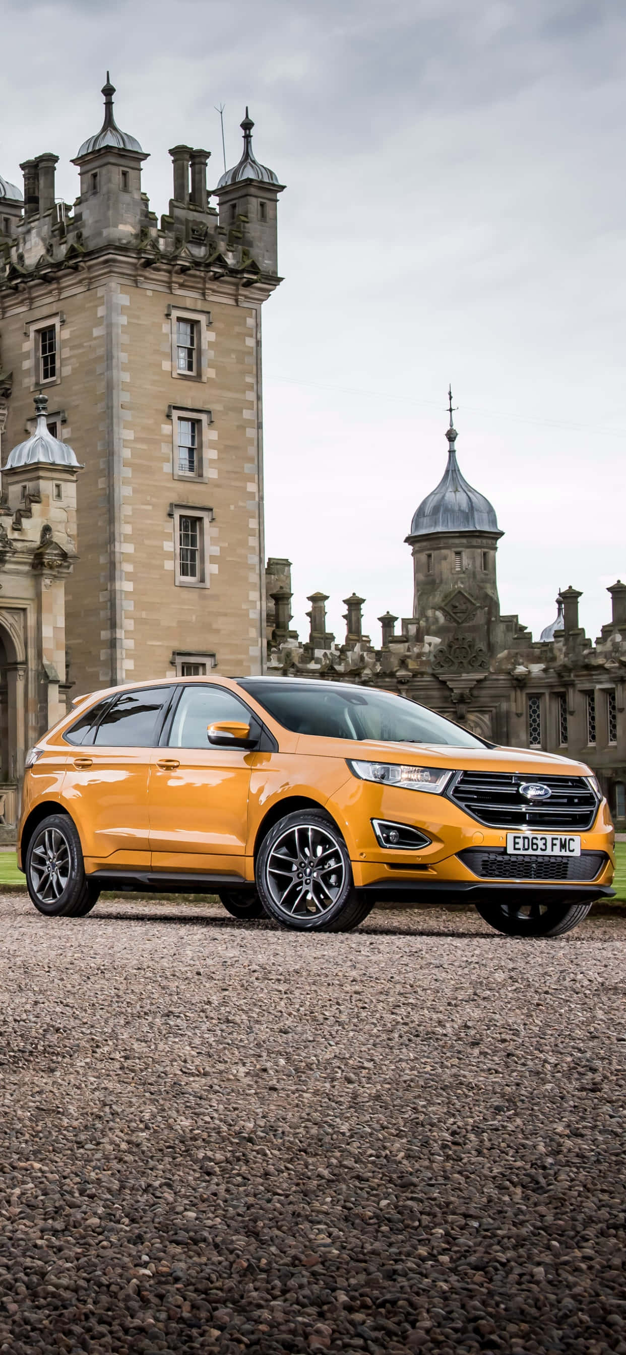 Ford Edge 桌布 壁紙 — 一輛鮮橙色SUV停泊在歷史石城堡前的碎石地上，天空多雲。