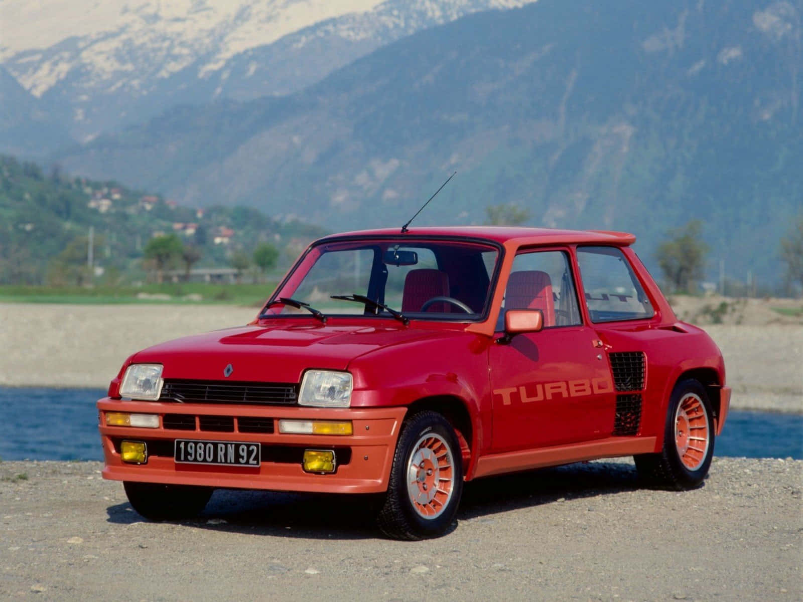 Renault 5 Turbo 桌布 壁紙 — 一輛鮮紅色的雷諾5 Turbo停泊在碎石上，旁邊是水，背景是白雪覆蓋的山脈。