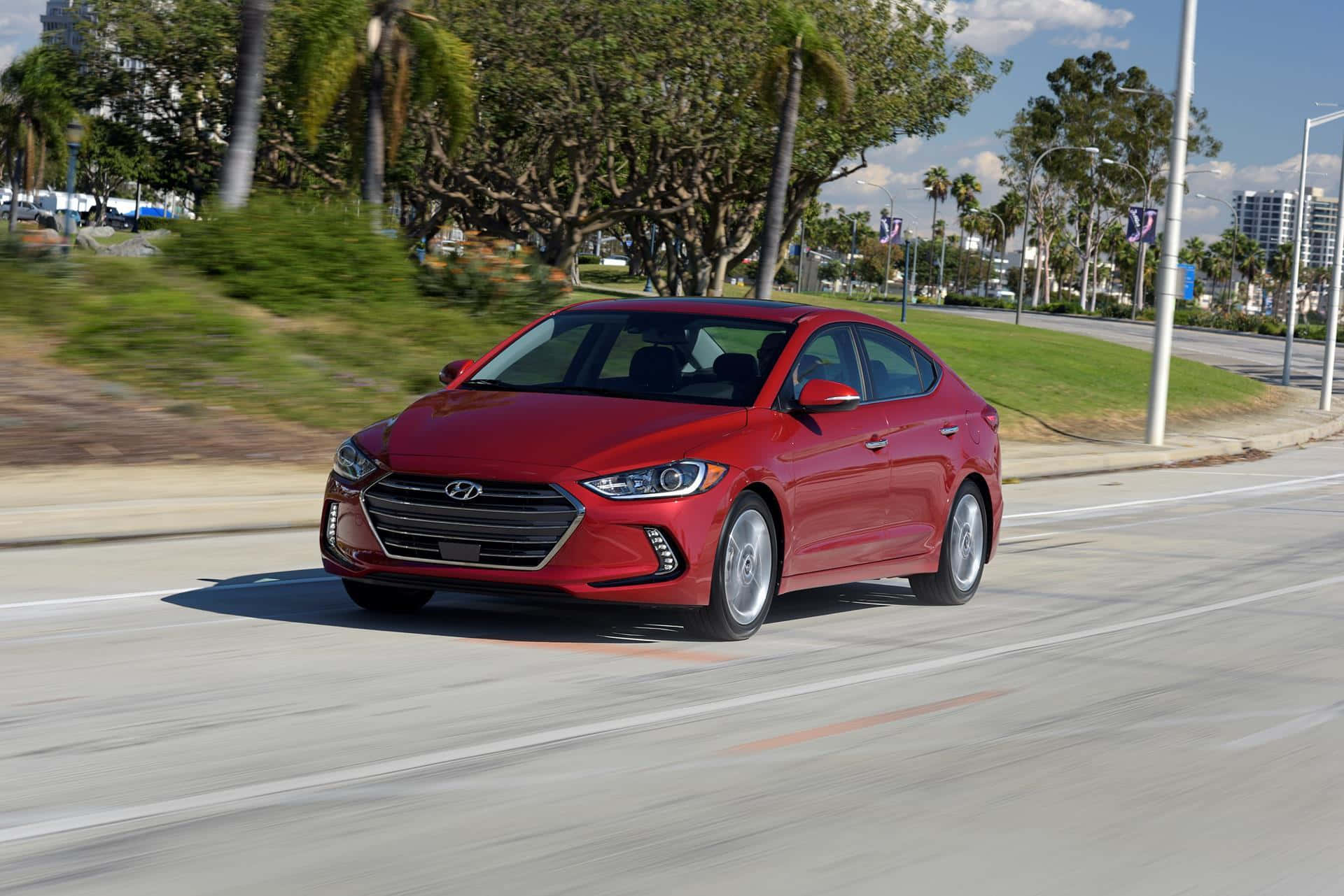 Hyundai Elantra 桌布 壁紙 — 一輛鮮紅色的現代房車在陽光明媚、兩旁種滿棕櫚樹和綠植的城市道路上飛馳。