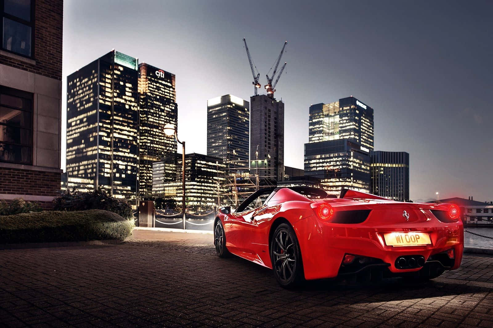 Ferrari 458 Spider 桌布 壁紙 — 一輛鮮紅色的法拉利跑車停泊在黃昏時分的鵝卵石街道上，背景是燈火通明的摩天大樓和起重機。