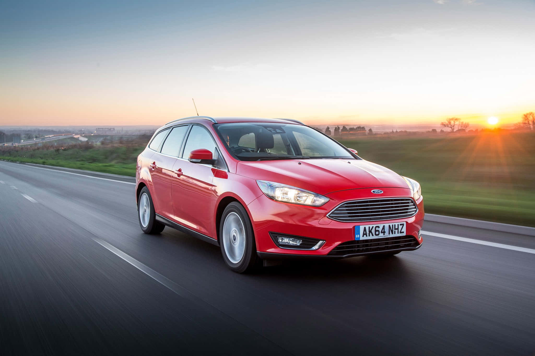 Ford Focus 桌布 壁紙 — 一輛鮮紅色的福特福克斯旅行車在日落時分的高速公路上飛馳，金色光芒在車後閃耀。