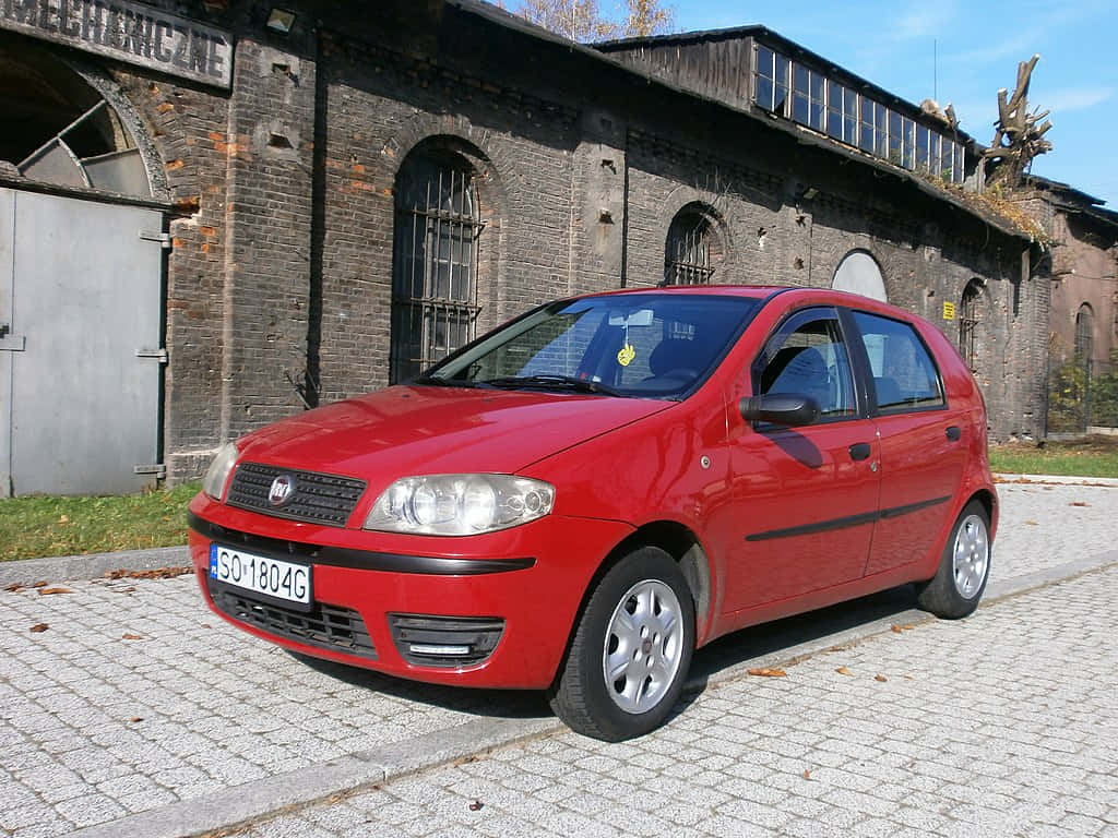 Fiat Punto 桌布 壁紙 — 一輛鮮紅色的快意Punto停泊在鵝卵石街道上，前方是一座風化的磚造工業建築，天空清澈湛藍。