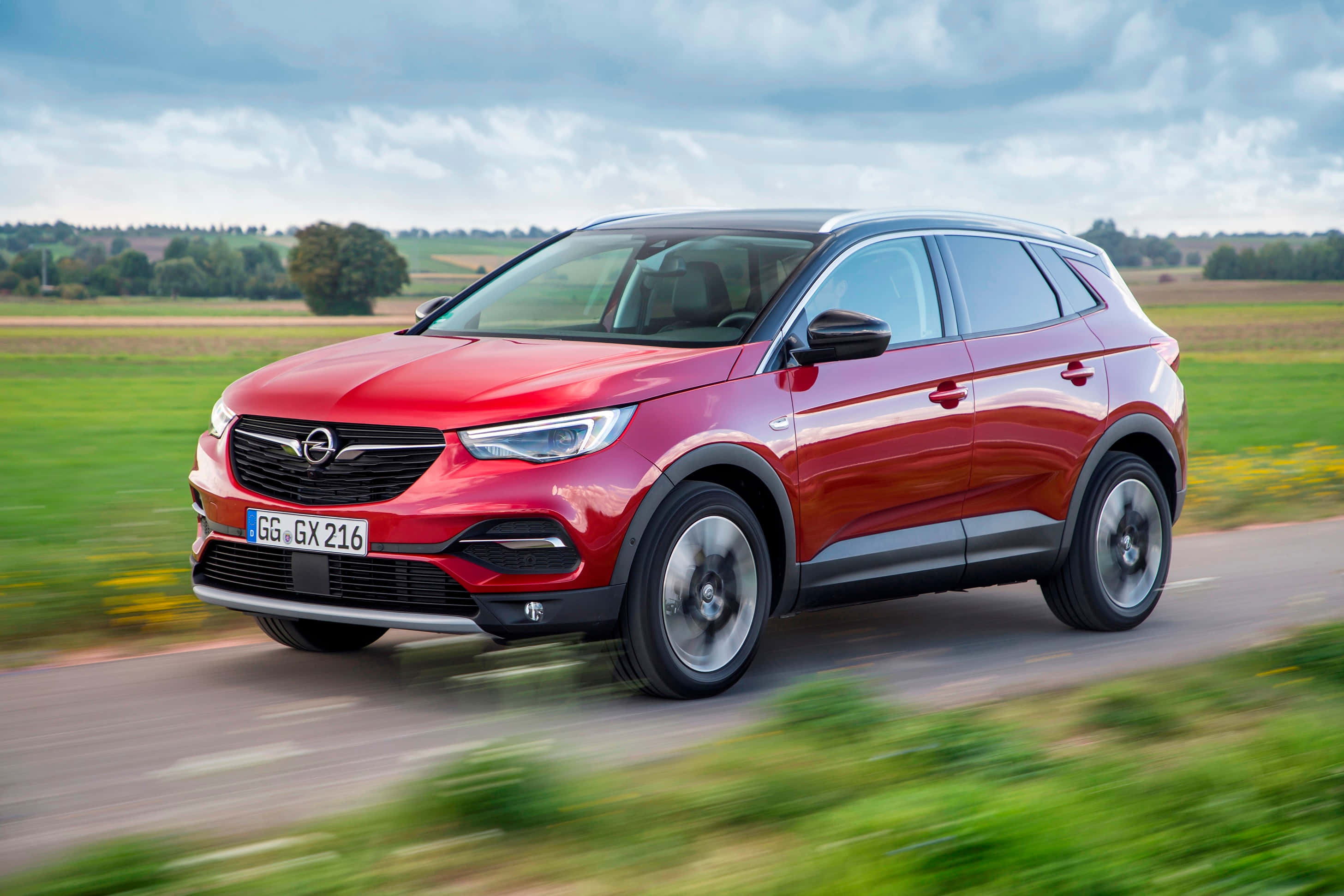 Opel Grandland X 桌布 壁紙 — 一輛鮮紅色的Opel SUV在鄉村道路上飛馳，背景是模糊的綠色田野和多雲的天空。