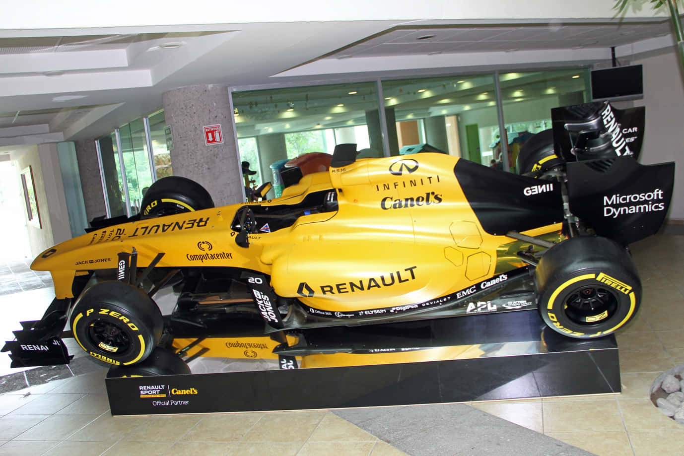 Renault Rs16 桌布 壁紙 — 一輛鮮黃色的雷諾一級方程式賽車，帶有黑色點綴和贊助商標誌，展示在現代室內大堂的基座上。