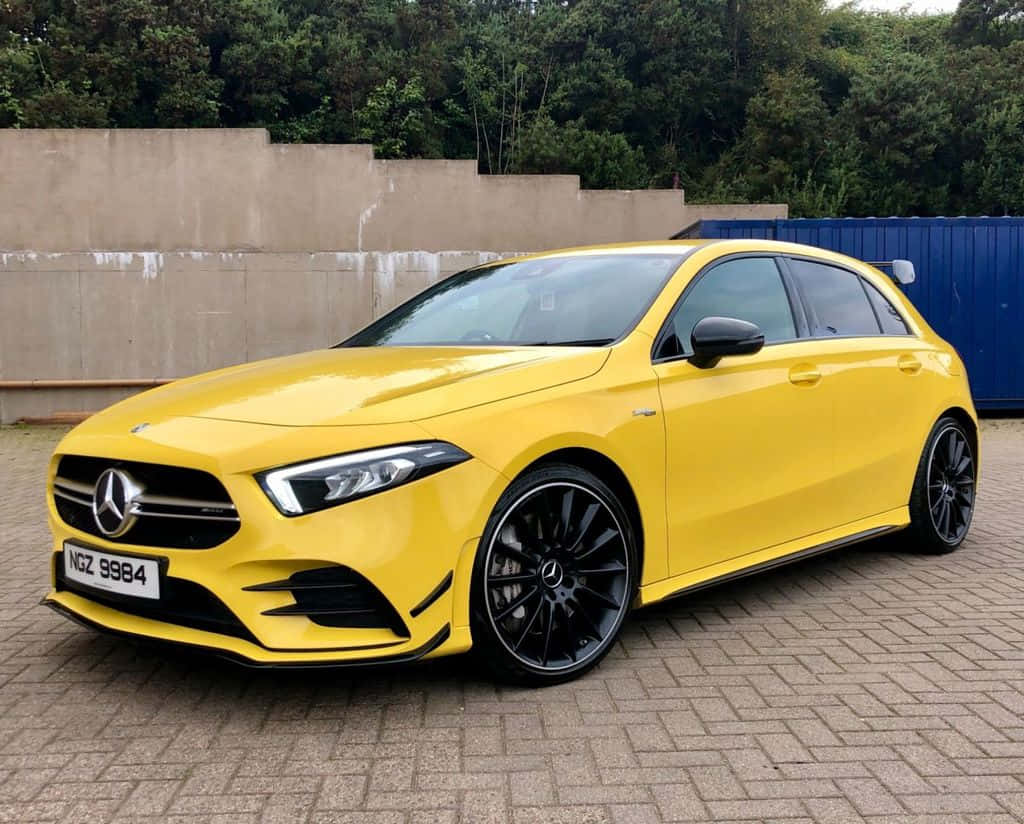 Mercedes Benz A-class 桌布 壁紙 — 一輛鮮黃色的Mercedes-AMG A45 S跑車停泊在鋪砌的停車場，背景有綠樹和混凝土牆。