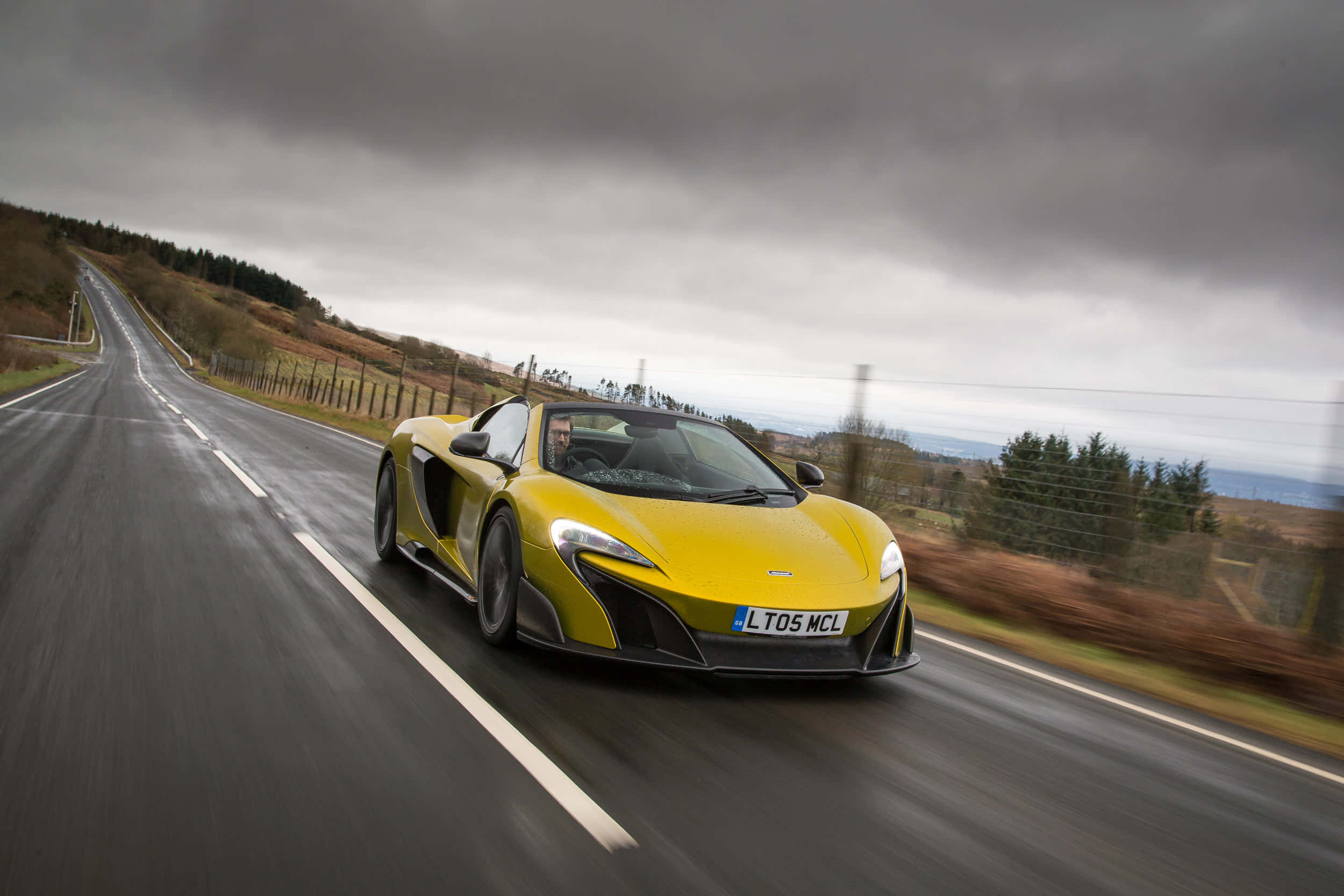 Mclaren 675lt Spider 桌布 壁紙 — 一輛鮮黃色的McLaren跑車在沉重的灰色天空下，沿著潮濕的鄉村道路飛馳。