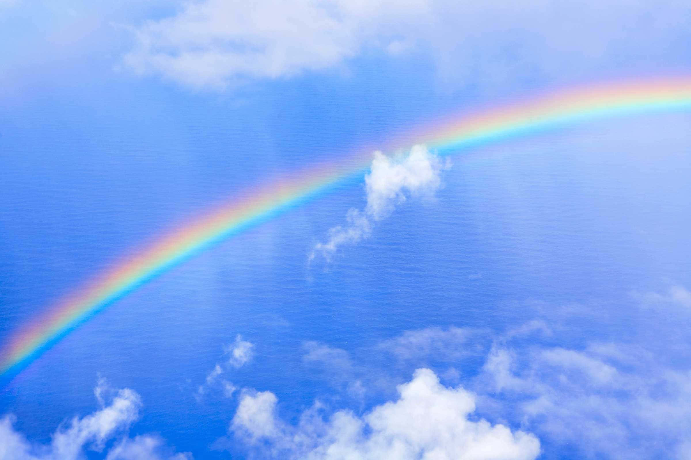 Rainbow Illusions 桌布 壁紙 — 一道鮮豔的彩虹優雅地拱起在寧靜的藍色海洋之上，天空部分多雲。