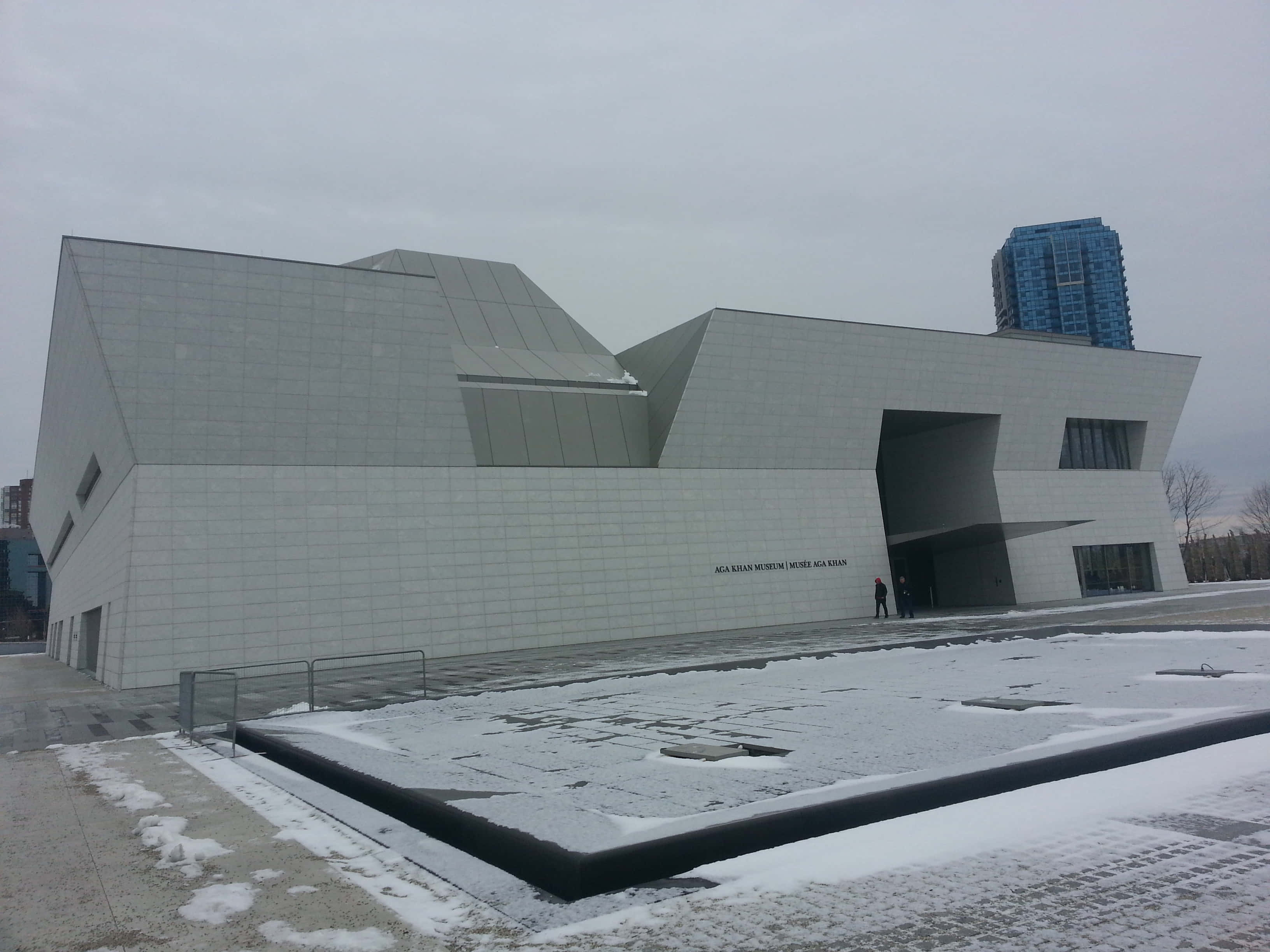 Aga Khan Museum 桌布 壁紙 — 一座現代幾何線條的博物館，白色瓷磚牆壁，矗立在雪覆蓋的城市廣場上，天空多雲。