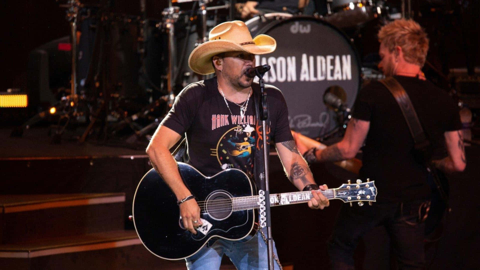  壁紙 — 鄉村歌手Jason Aldean在舞台上表演，戴著牛仔帽，手持木結他，在舞台燈光下。