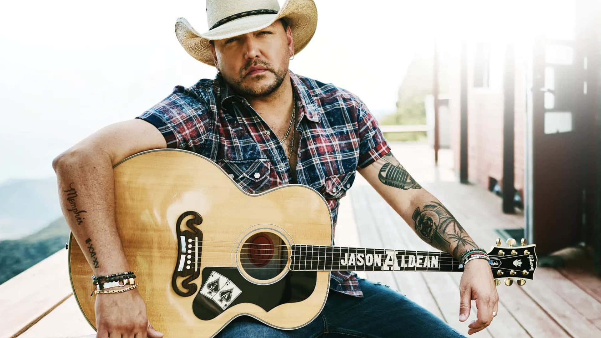  壁紙 — 鄉村歌手Jason Aldean坐在木質門廊上，手持一把琴頭刻有他名字的原聲結他。