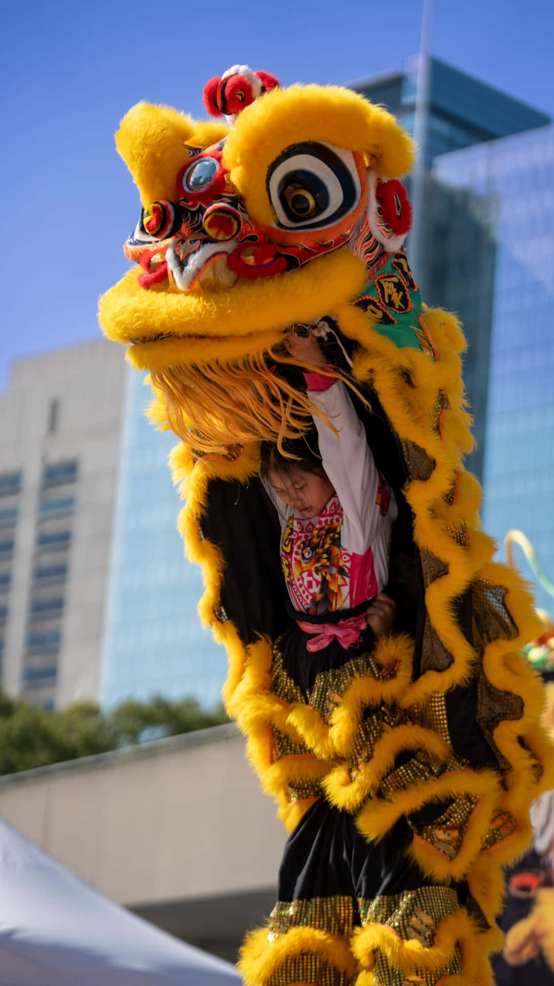  壁紙 — 一件鮮黃色的舞獅服裝，帶有精緻的紅色和金色細節，由一個人在現代都市環境中，在晴朗的藍天下表演。