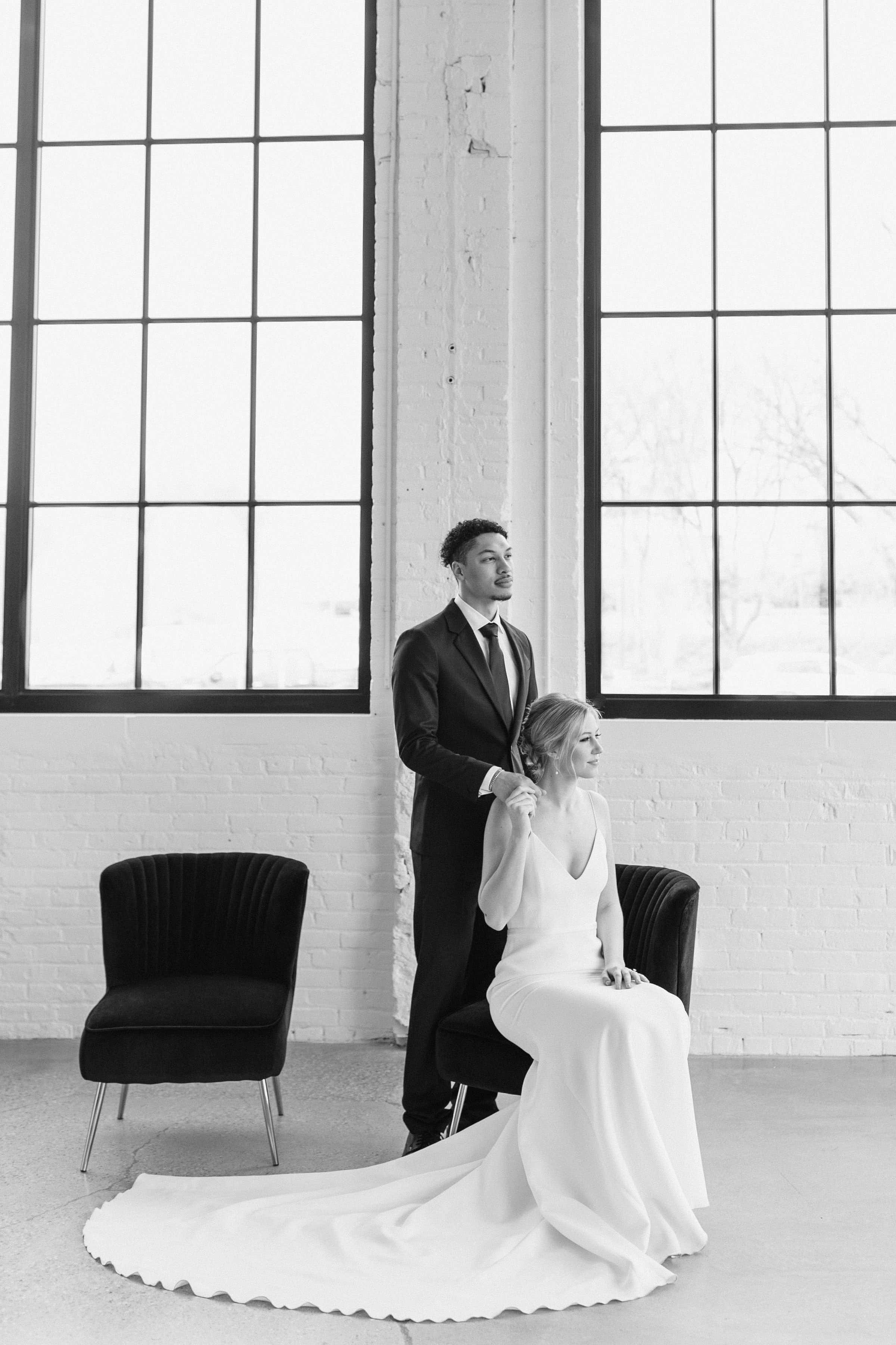 Black And White Wedding 桌布 壁紙 — 一位身穿飄逸白色婚紗的新娘優雅地坐在黑色椅子上，旁邊是身穿西裝的新郎，背景是極簡黑白工作室的大型工業窗戶。