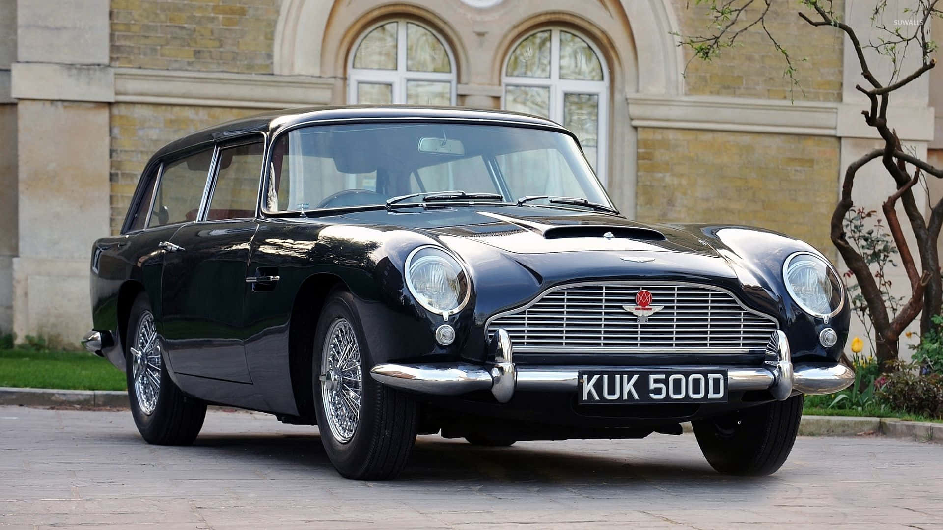 Aston Martin Db5 桌布 壁紙 — 一架型格黑色Aston Martin DB6 Shooting Brake停泊在石頭車道上，前方是古典石建築，帶有拱形窗戶。