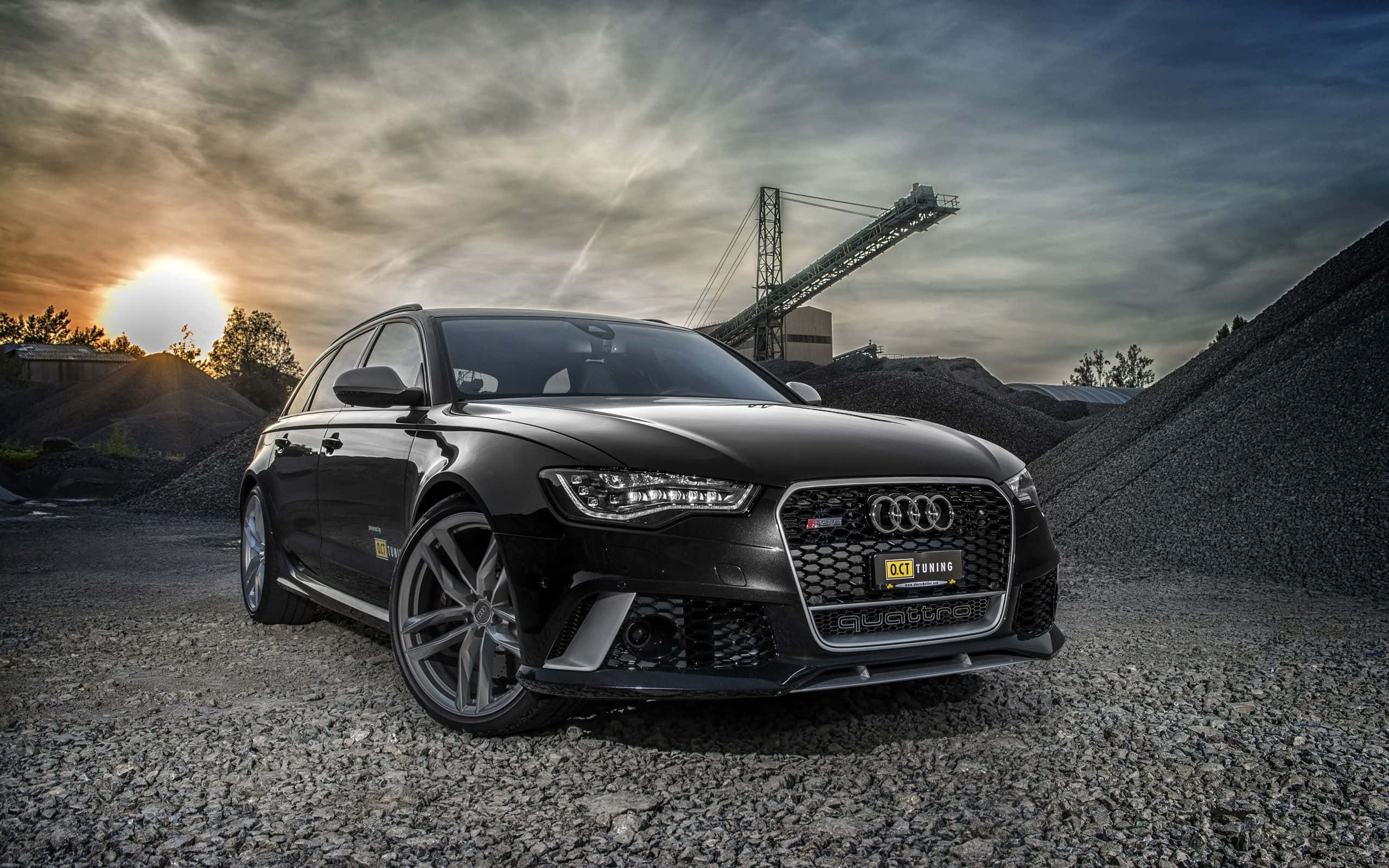 Audi S6 桌布 壁紙 — 一架型格黑色Audi RS6 Avant停泊在採石場的碎石上，背景有戲劇性的日落和工業輸送帶結構。