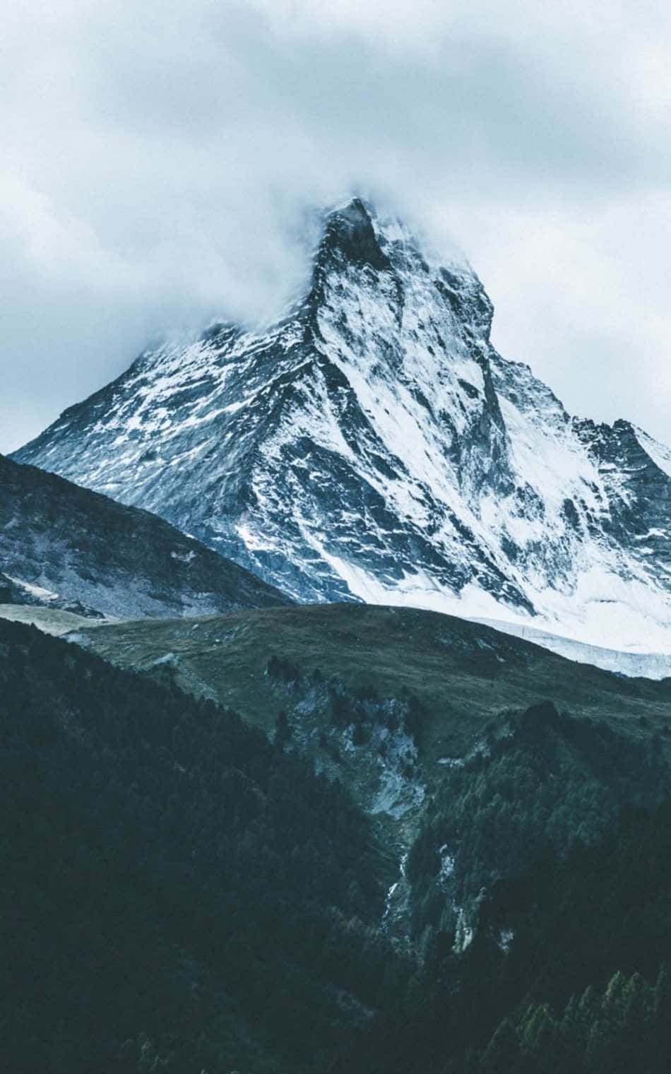  壁紙 — 在灰色天空下，一座雄偉的雪山峰穿過低垂的雲層，聳立在森林覆蓋的山谷之上。