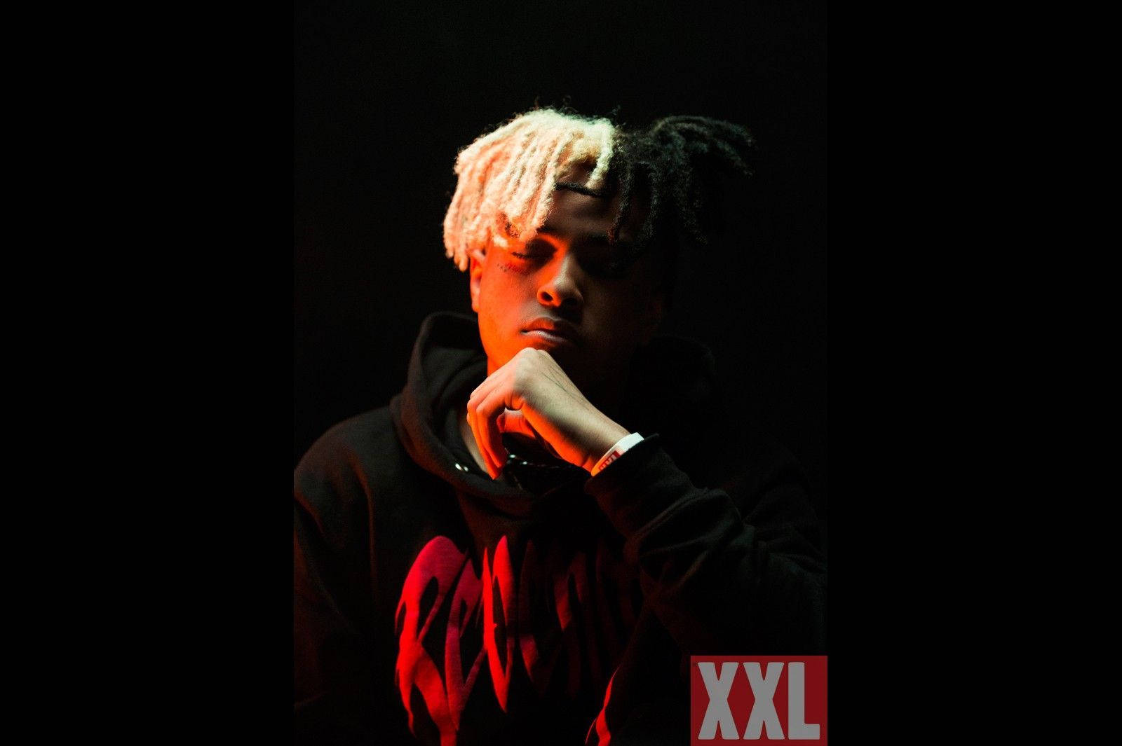 XX Tentacion 深色背景肖像 壁紙 — 一位擁有雙色辮子髮型的年輕男子，在黑色背景前，於戲劇性的紅光下將下巴擱在手上。
