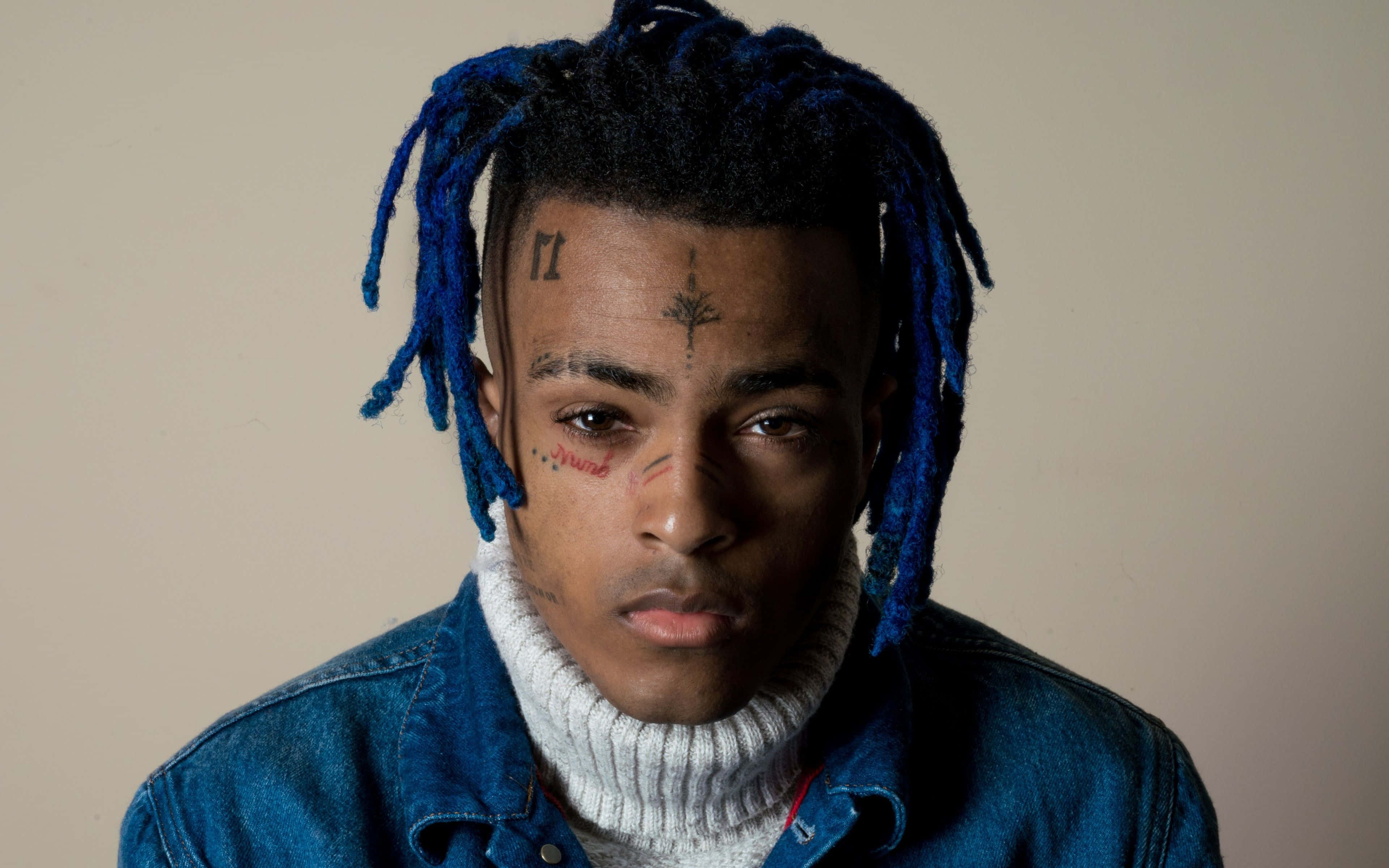 XXXTentacion 坐在筆記本電腦前，周圍環繞書籍與藝術用品 壁紙 — 一名男子嘅特寫肖像，佢有鮮豔嘅藍色辮子同面部紋身，著住牛仔褸同白色高領衫。