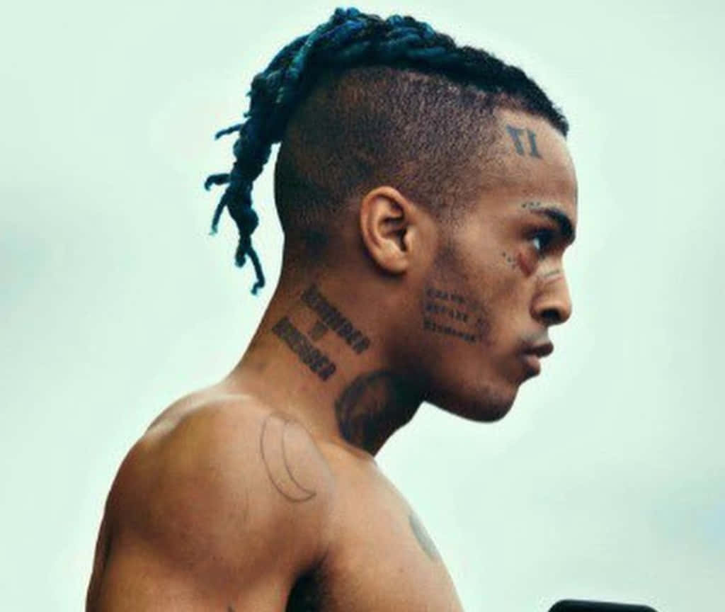 Xxxtentacion 側面紋身頭像 壁紙 — 一個有藍色辮子頭和頸部及面部紋身的男人，在純色背景下強烈地凝視。