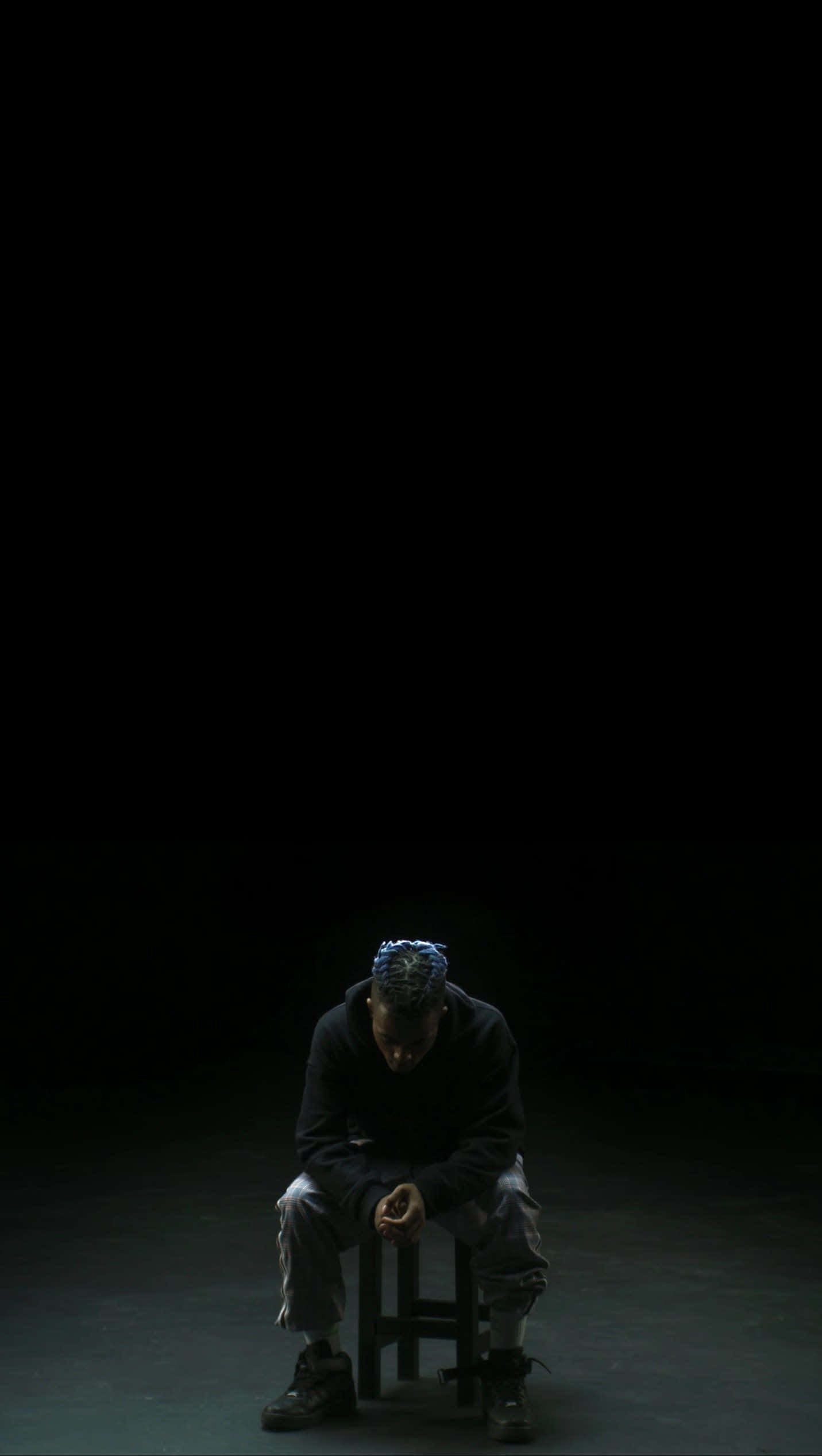 藍髮XXXTentacion Bad 壁紙 — 一個孤獨的人蜷縮在凳子上，身處漆黑虛空，被單一聚光燈照亮，投射出戲劇性的陰影。