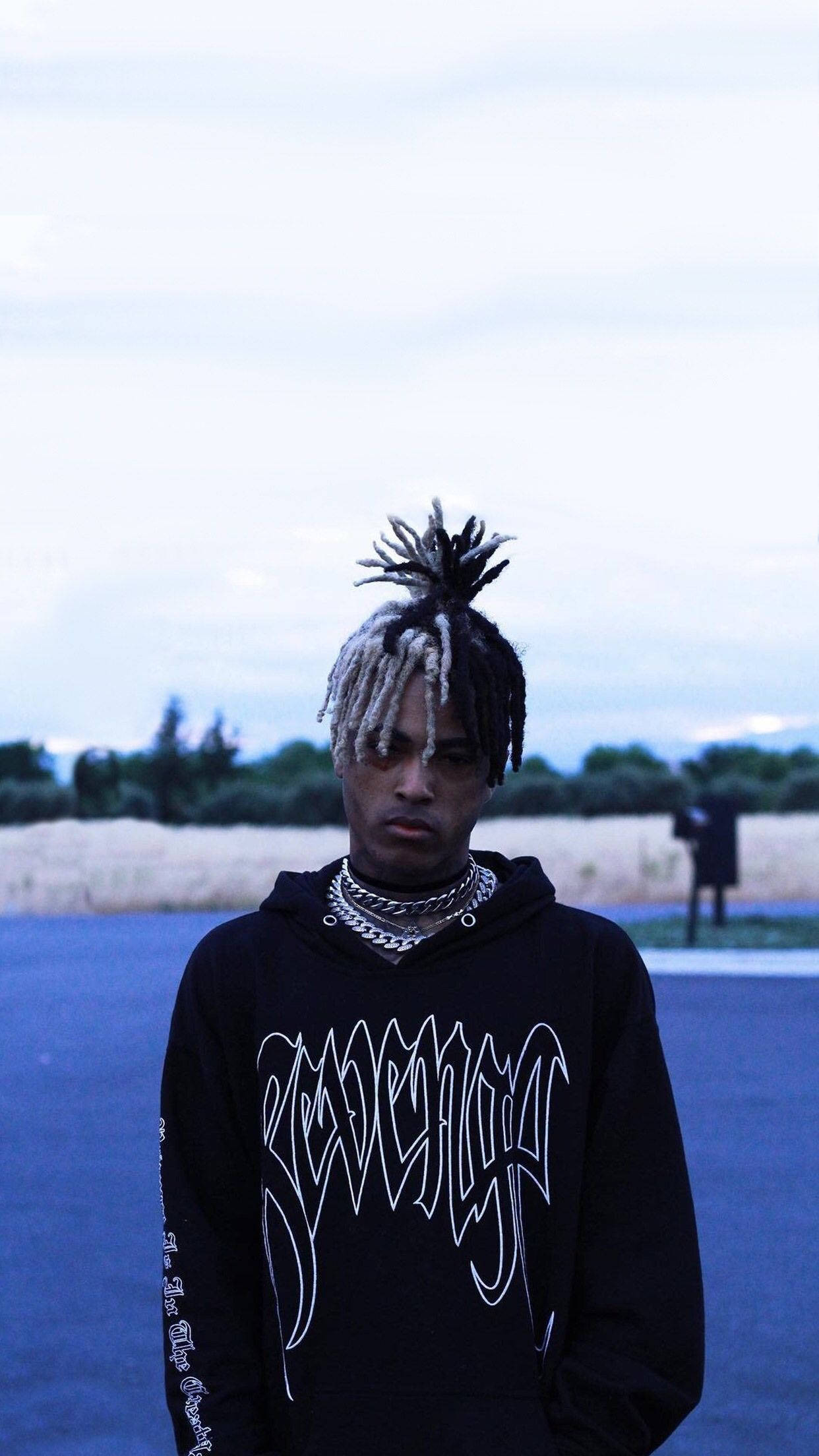 XXXTENTACION粉絲身穿其標誌性黑色連帽衫 壁紙 — 一位年輕男子編著辮子，戴著多層鏈條，在陰天、冷色調的戶外環境中自信地站立。