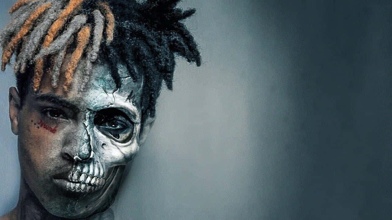 XXXTentacion 的創意筆記本電腦 壁紙 — 一個人半邊臉是骷髏半邊是人類，帶有橙色和黑色的辮子，背景為柔和的灰色。
