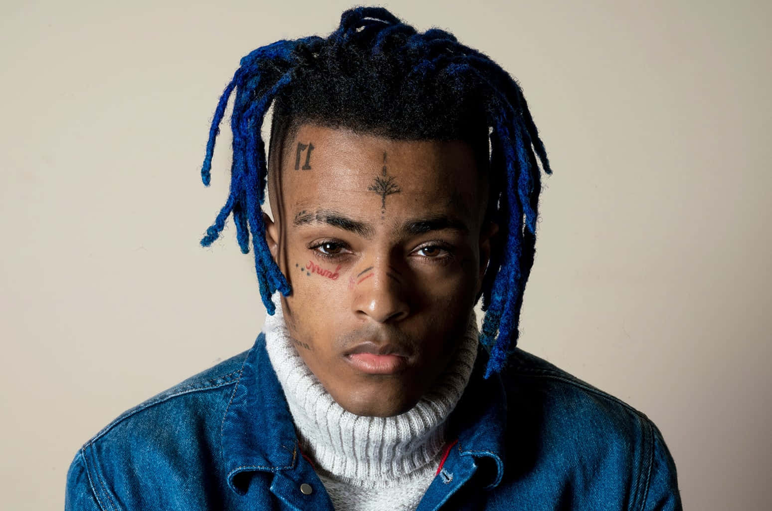 藍色捲髮已故XXXTentacion 壁紙 — 一位年輕男子擁有充滿活力的藍色辮子頭和臉部紋身，在純米色背景前凝視著相機。
