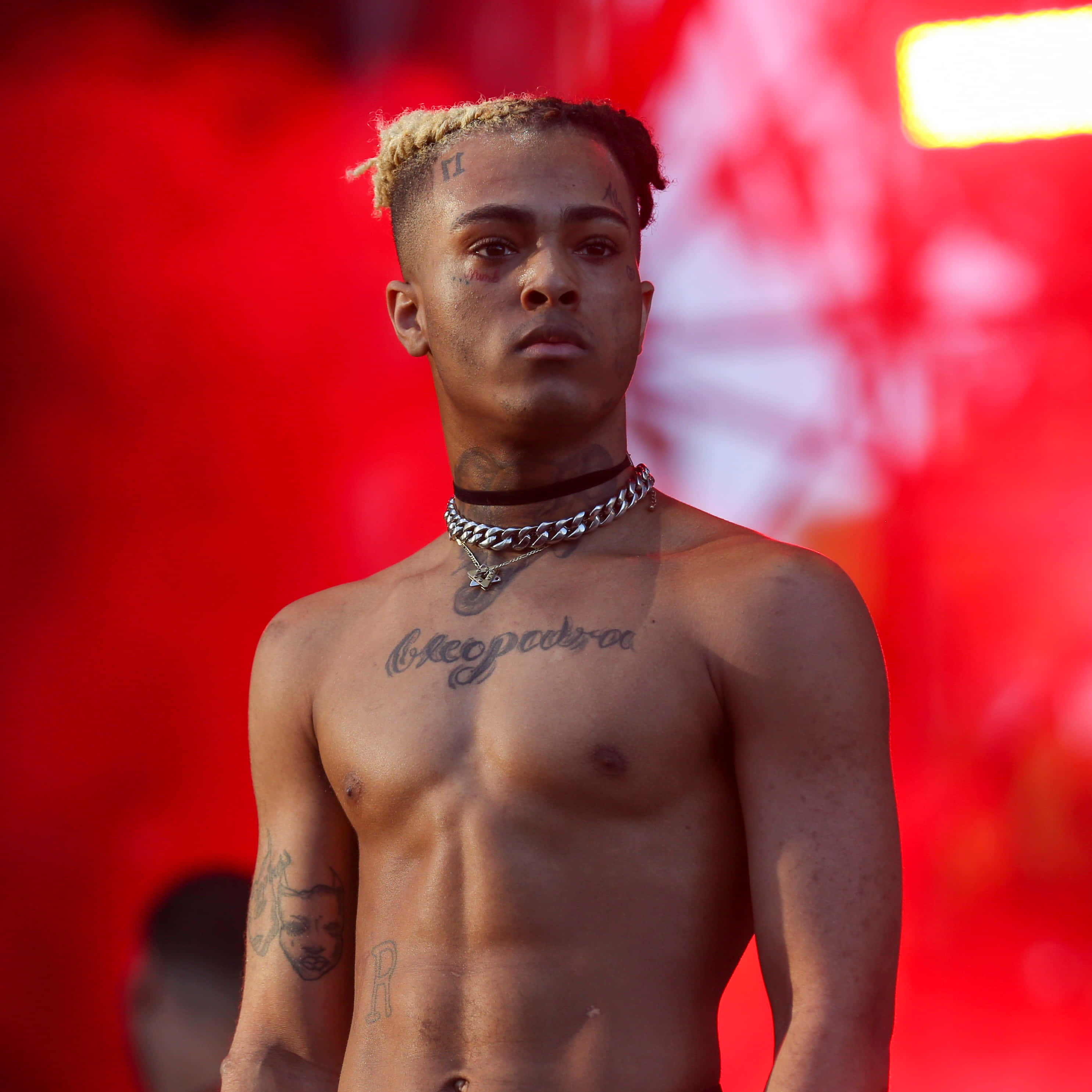 Xxxtentacion 赤裸上身頭像 壁紙 — 一個赤裸上身的男人，金色辮子和胸口紋身，在舞台燈光下站在鮮紅色背景前。
