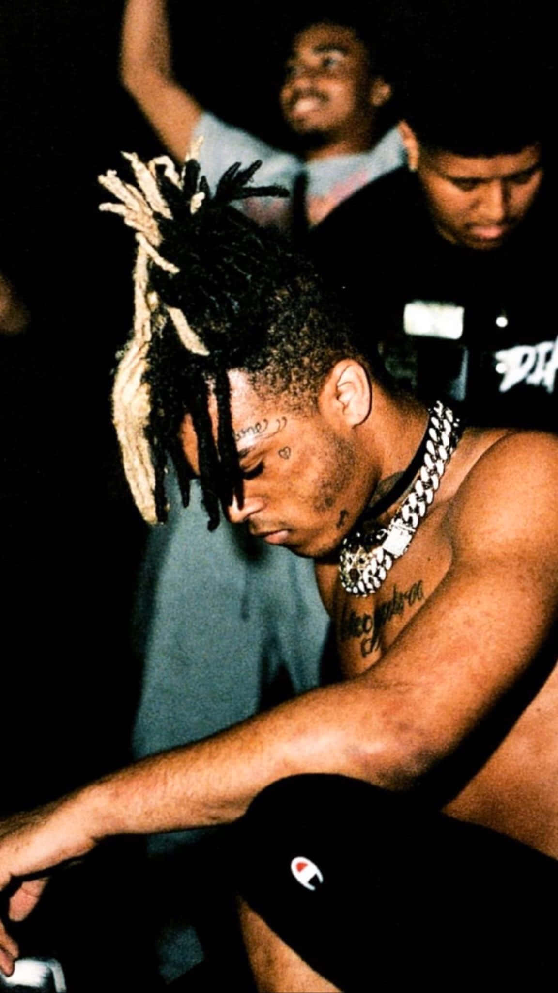 Xxxtentacion 跪姿無上衣頭像 壁紙 — 一個冇著衫、有辮子頭同紋身嘅男人喺昏暗光線下坐低，戴住一條粗鏈，而其他人企喺佢後面。