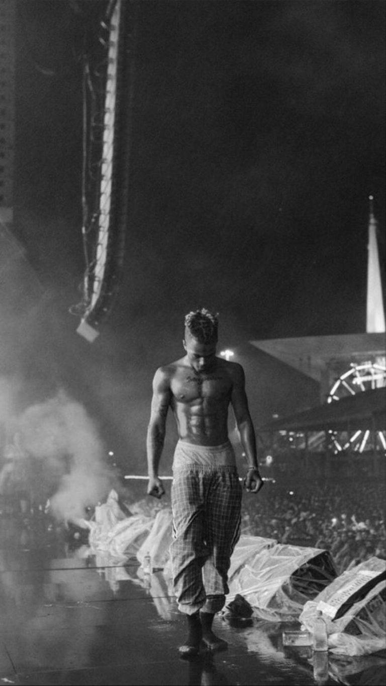 XXXTentacion 演唱會黑白美學 壁紙 — 一個赤膊肌肉男赤腳走過煙霧，在夜燈下的濕滑舞台上，下方有群眾。