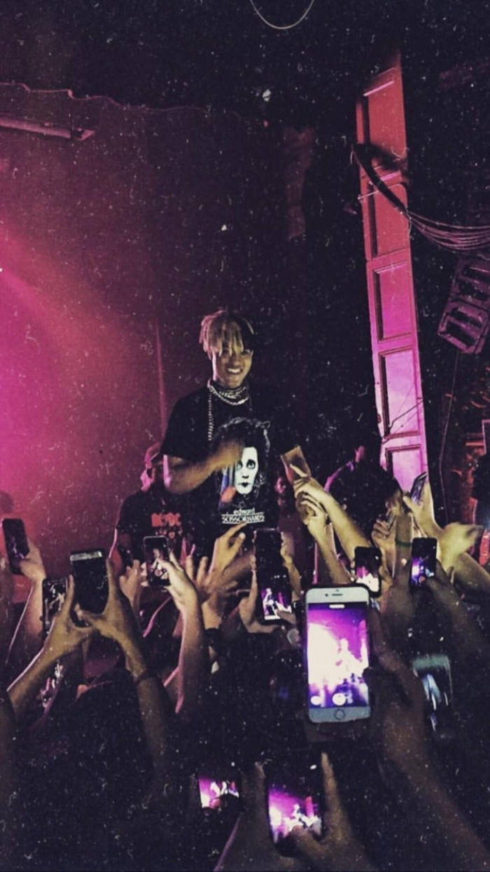 XXXTentacion 演唱會親密互動 壁紙 — 一位戴著白色面具的表演者在粉紅色舞台燈光下，在一群高舉發光手機的粉絲上方咧嘴而笑。
