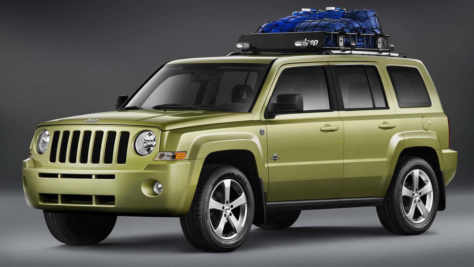 Jeep Patriot 桌布 壁紙 — 一架啞光橄欖綠色的Jeep Patriot SUV，車頂架上有一個藍色貨物箱，站在深灰色工作室背景前。