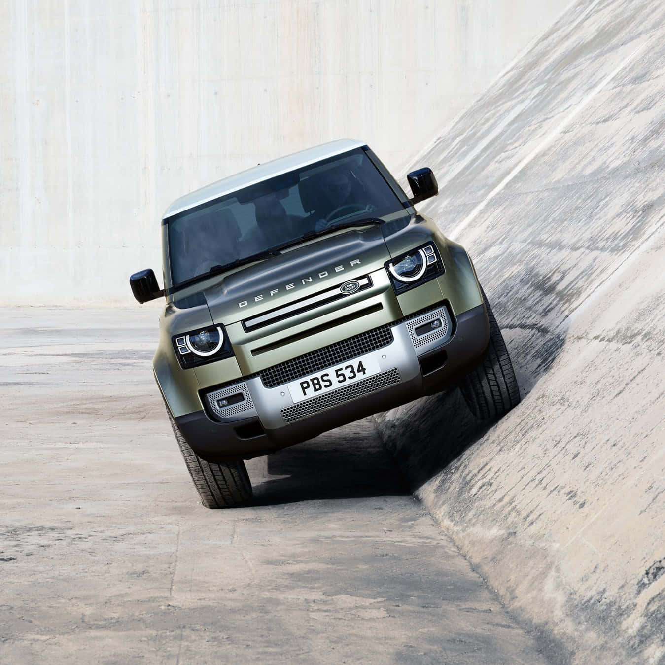 Land Rover Defender 桌布 壁紙 — 一輛啞光橄欖綠色的 Land Rover Defender 在簡樸的工業環境中急劇地傾斜在混凝土斜坡上。