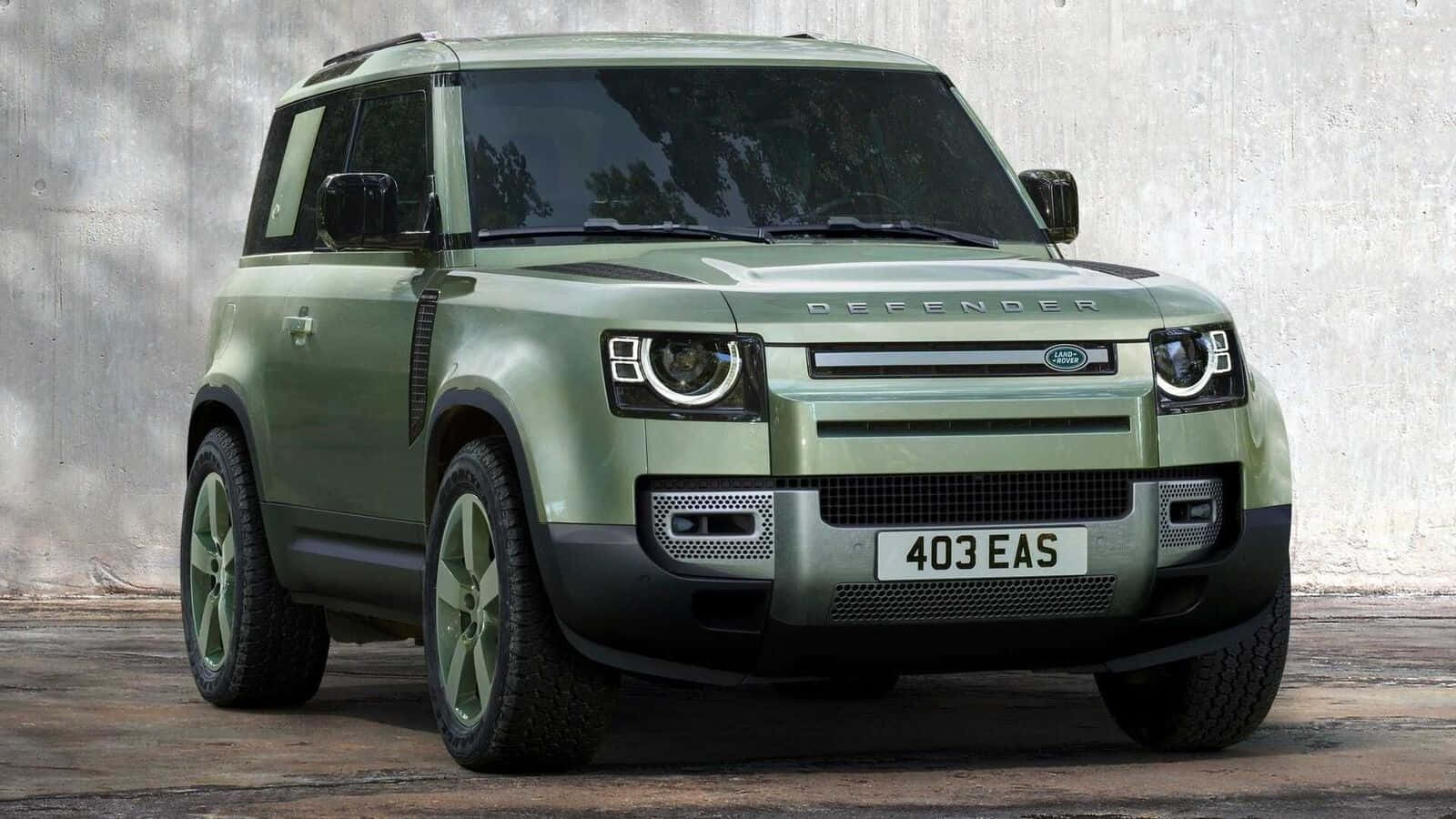 Land Rover Defender 桌布 壁紙 — 一輛啞光橄欖綠色的Land Rover Defender在明亮的日光下，大膽地停在一面風化的混凝土牆前。