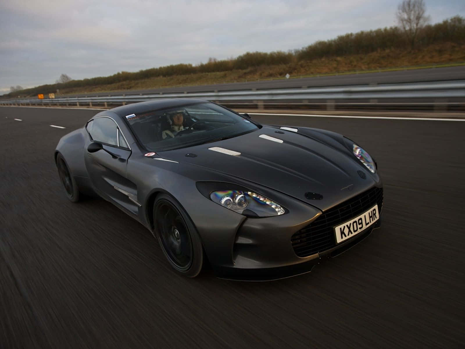 Aston Martin One-77 桌布 壁紙 — 一輛啞光黑色的Aston Martin V12 Vantage在高速公路上奔馳，帶有動態模糊和陰天。