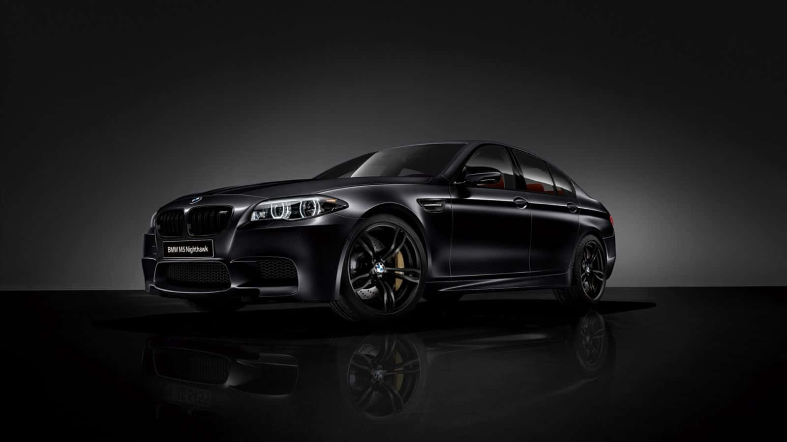 Bmw M5 桌布 壁紙 — 一輛時尚的啞光黑色BMW M5 Nightshade轎車在攝影棚燈光下閃耀，金色制動卡鉗在深色漸變背景上清晰可見。