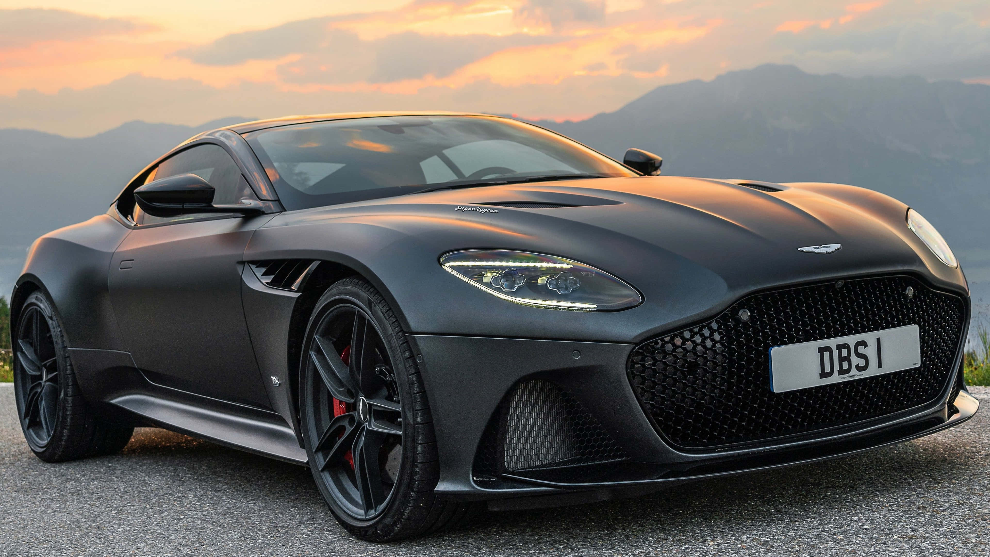 Aston Martin Dbs Superleggera 桌布 壁紙 — 一輛啞光黑色的Aston Martin DBS跑車在金色日落和山脈背景下閃閃發光。