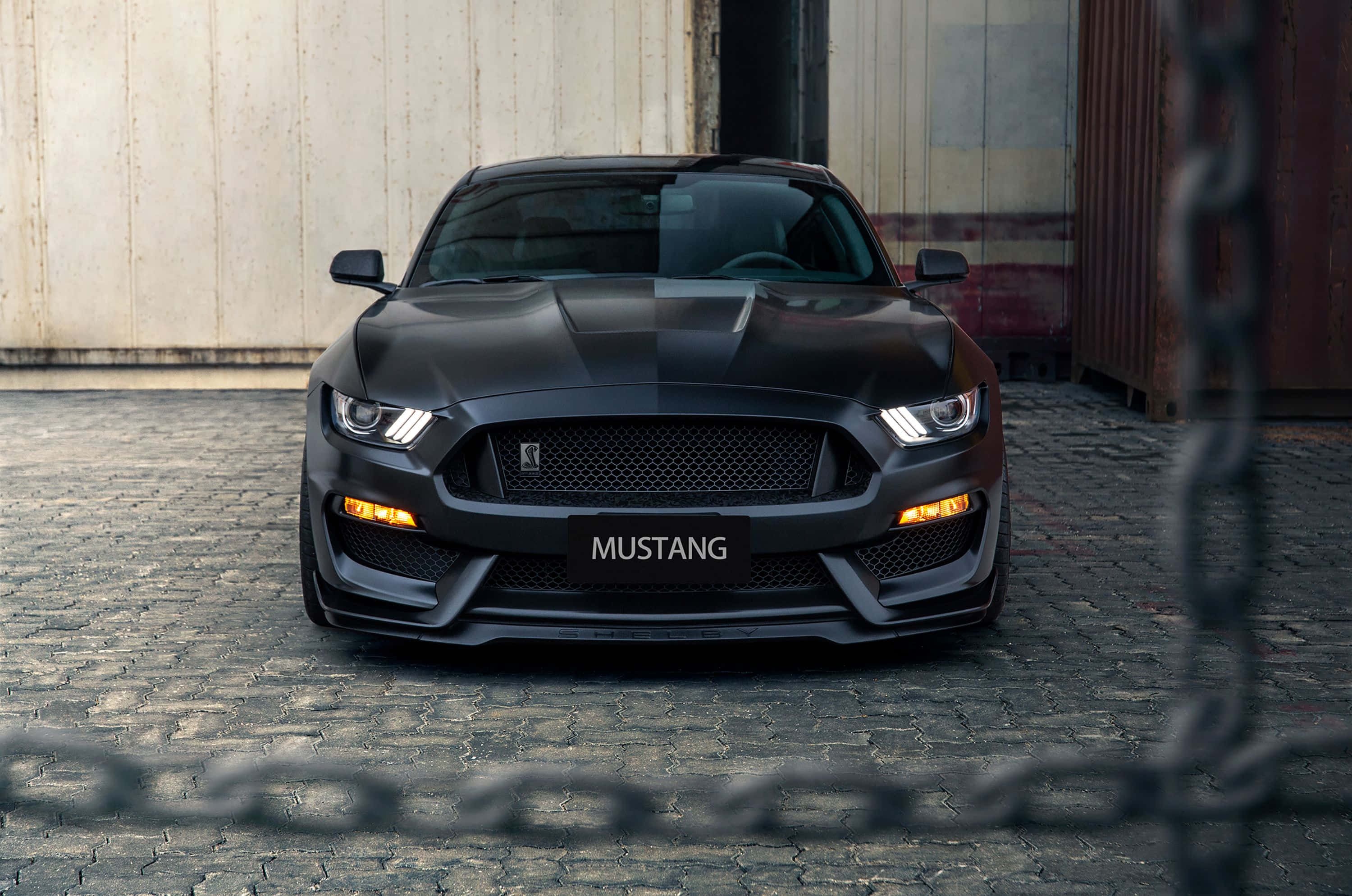 Ford Mustang Shelby Gt350 桌布 壁紙 — 一輛時尚的啞光黑色福特野馬跑車，車頭燈發光，停在工業集裝箱前的鵝卵石街道上。