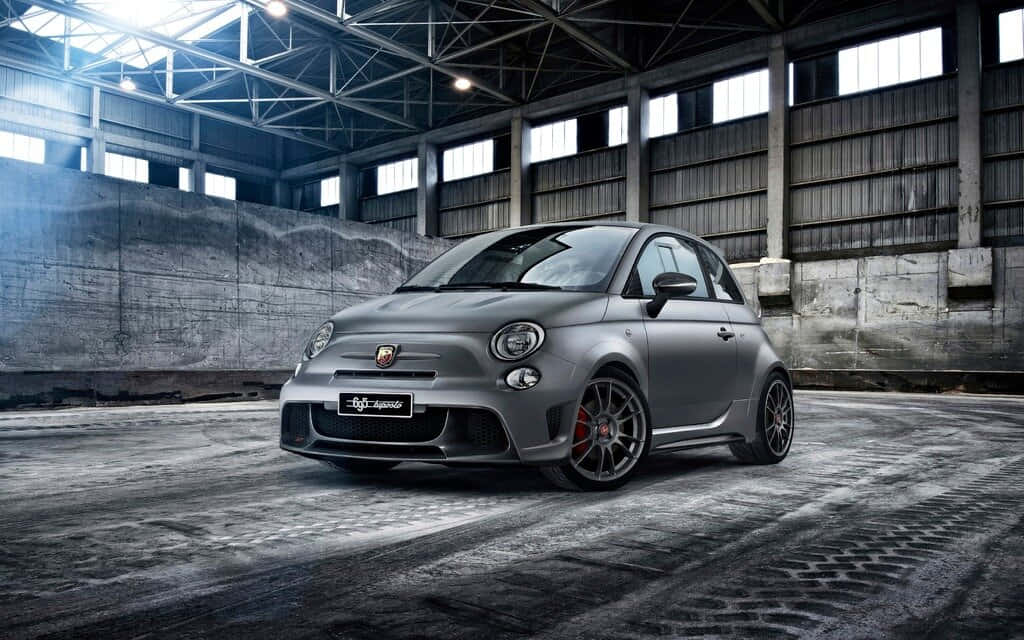 Fiat 500 桌布 壁紙 — 一輛啞光灰色的Fiat 500 Abarth跑車，帶有紅色制動卡鉗，停在一個粗糙的工業倉庫中，天花板很高，地板是混凝土的。